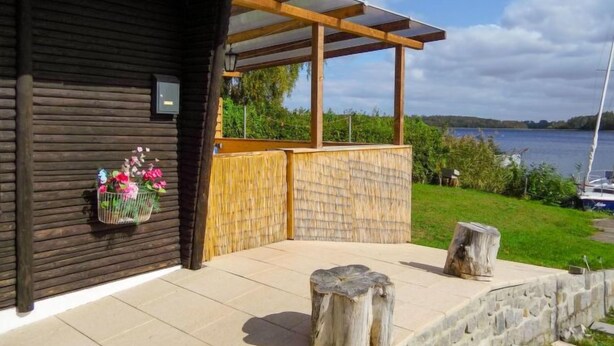 A wooden cabin with a covered patio, a stone wall, and a view of a lake. - Güstrower Chaussee by Interhome (Krakow am See)