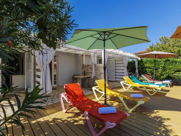 A wooden deck with sun loungers and umbrellas, a stone wall, and a covered seating area with a table and chairs.
