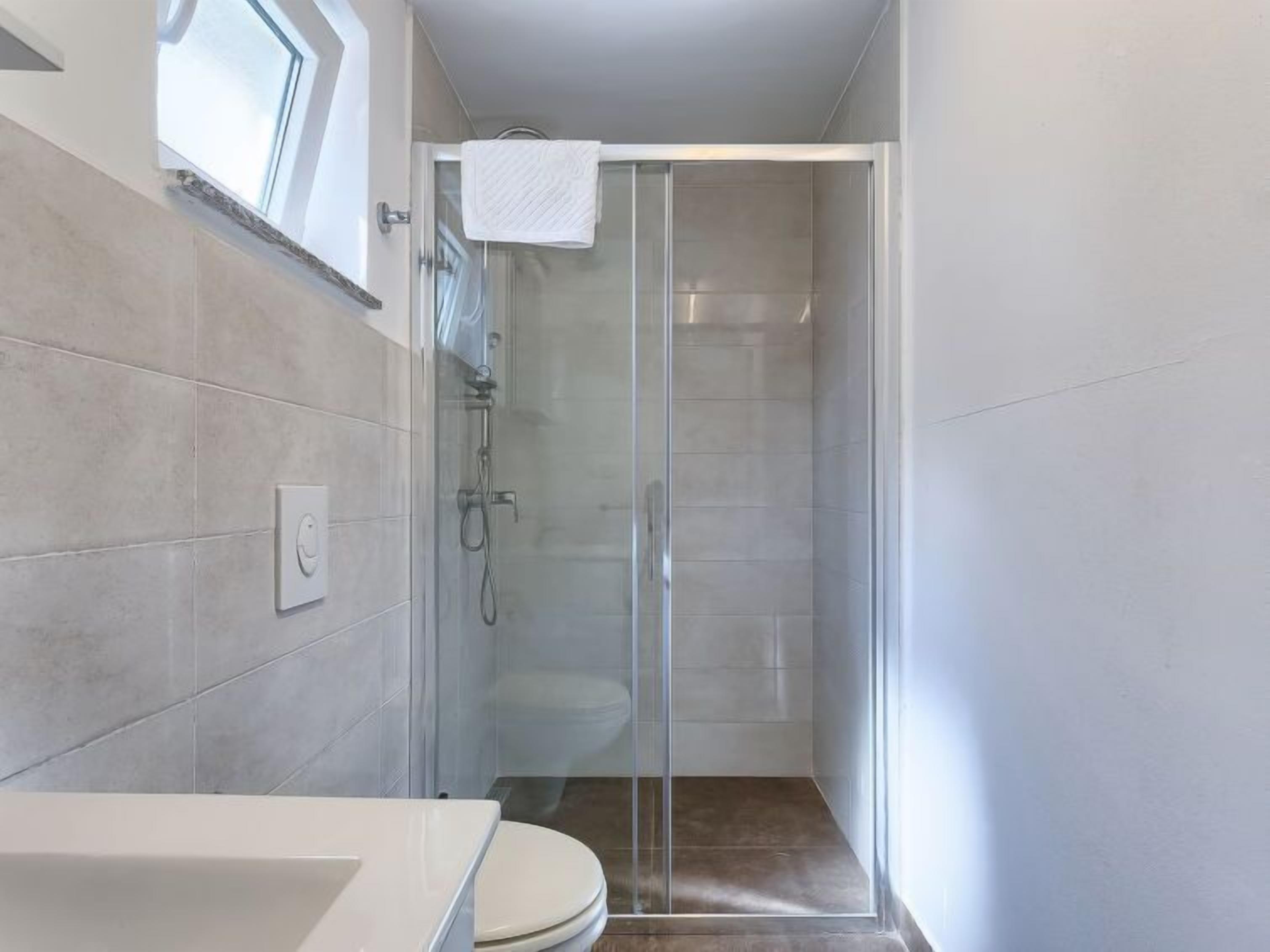 A modern bathroom with a glass shower enclosure, a white toilet, and a window above the sink.