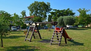 Un'area giochi con altalene e scivolo, immersa nel verde con alberi e auto parcheggiate sullo sfondo.