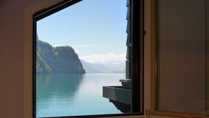 Ein Zimmer mit großem Fenster und Blick auf einen stillen See sowie eine gebirgige Landschaft.