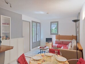 Une pièce compacte comprenant une table à manger pour deux personnes, un lit avec une tête de lit en bois, une petite télévision et une fenêtre avec des rideaux.