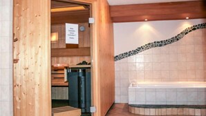 Un sauna avec des murs en bois, un banc, un banc avec une serviette, un radiateur noir et un mur recouvert de carreaux avec une bordure en mosaïque.