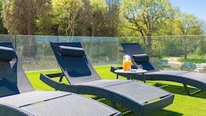 A sunlit outdoor lounge area with reclining chairs and a small table, surrounded by a glass fence and lush greenery.