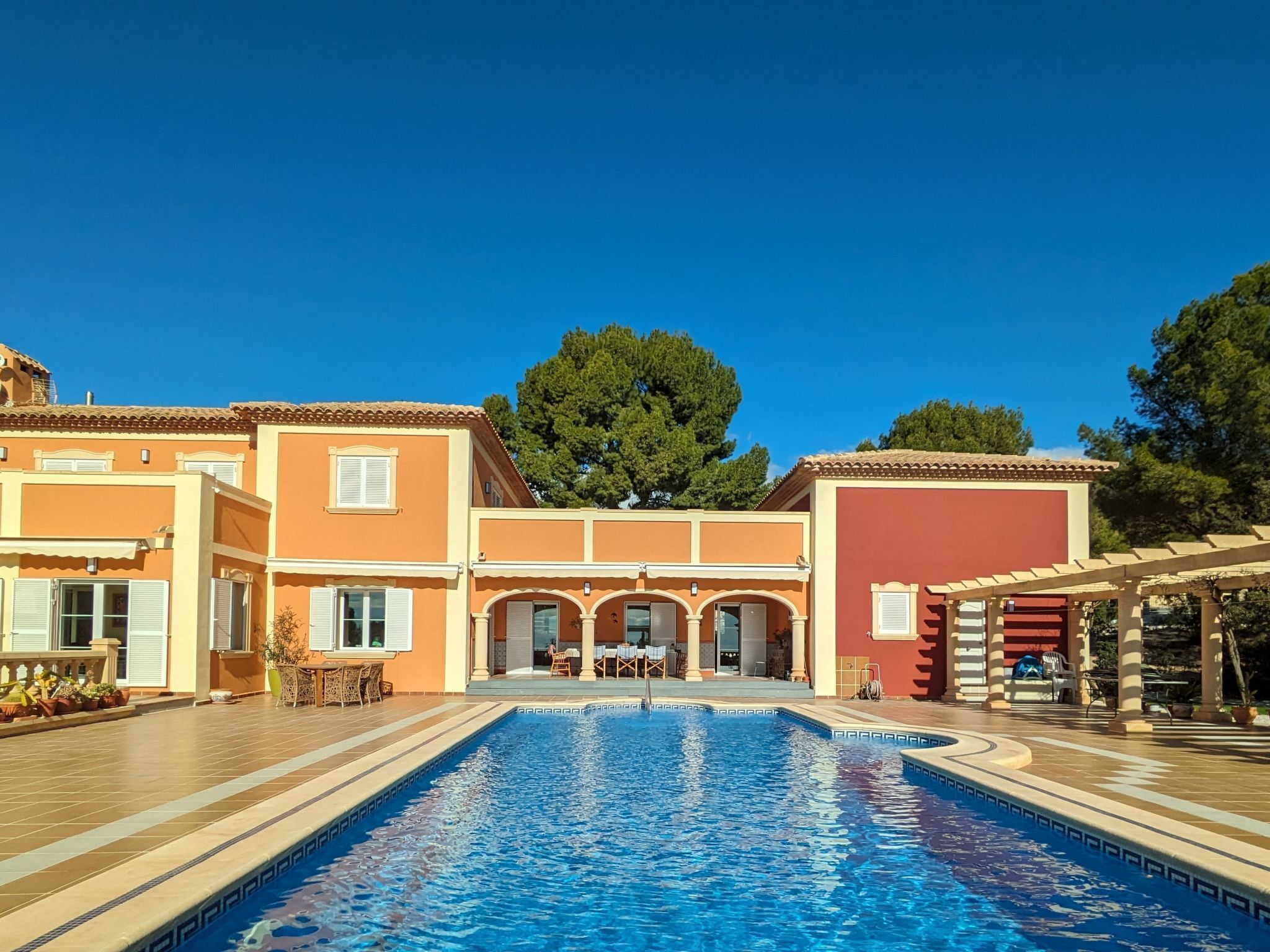 A swimming pool with a tiled deck, a covered seating area, and a building with multiple windows and a tree in the background.