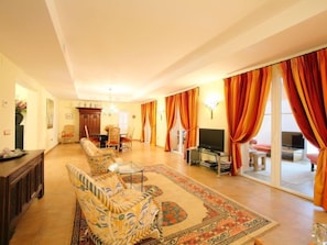 A spacious living room with a sofa, a coffee table, and a TV. There is a dining area with chairs and a table. The room has large windows with orange curtains.