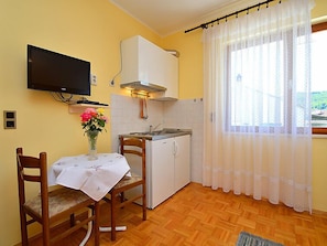 A compact kitchen with a dining table, chairs, a TV mounted on the wall, a small refrigerator, and a window with sheer curtains.
