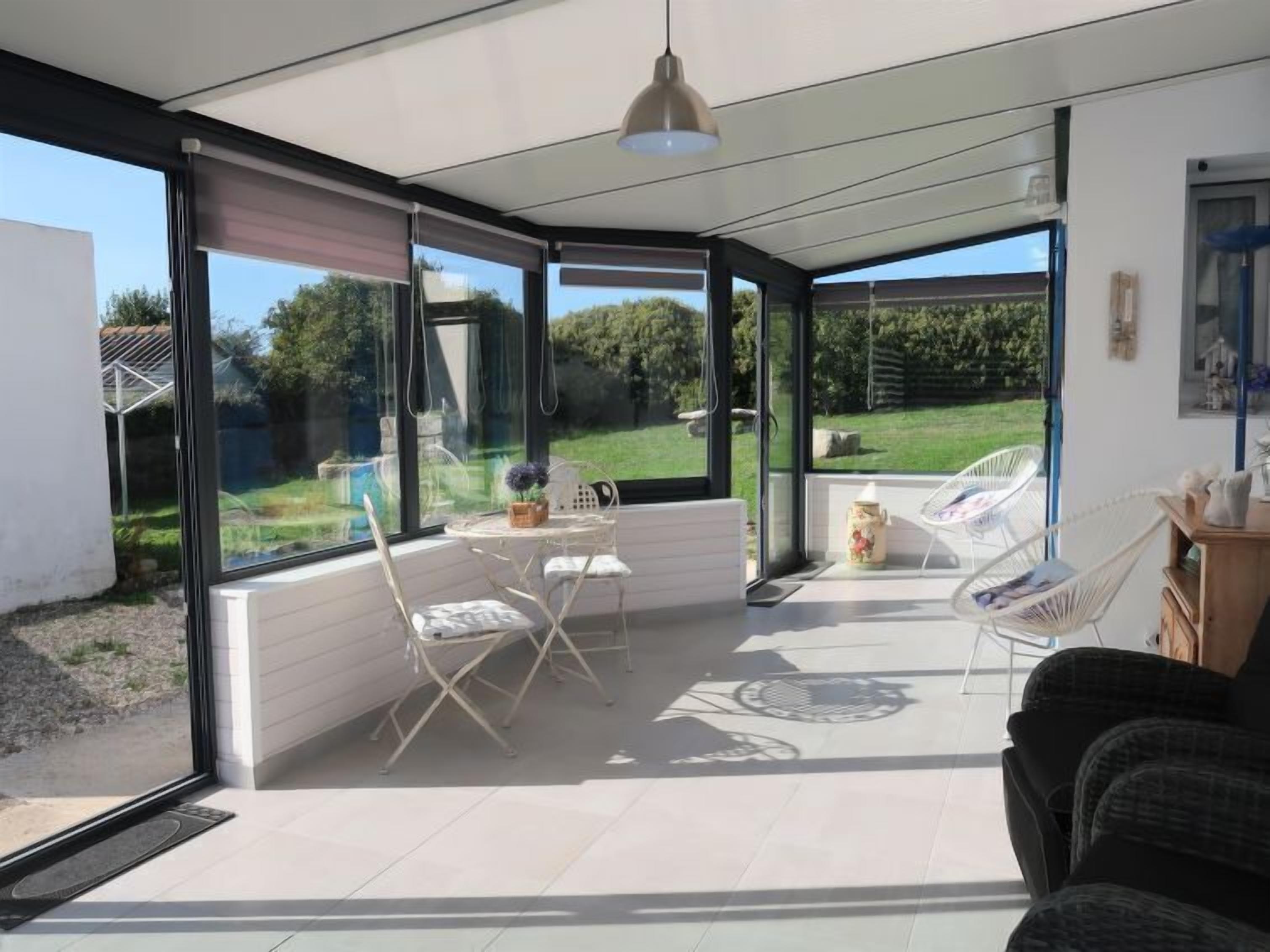 A modern, open-plan living space with large glass doors leading to a garden. There are white chairs and a small table with a vase.
