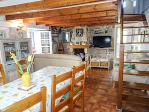 Un salon chaleureux avec une cheminée, des poutres apparentes, un coin repas avec une table et des chaises, et un téléviseur fixé au mur.
