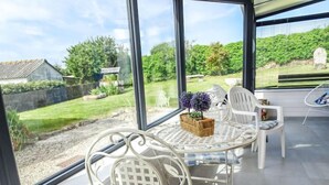 Een zonnig terras met een witte metalen tafel en stoelen, een rieten mand met bloemen en uitzicht op een tuin en een stenen structuur.