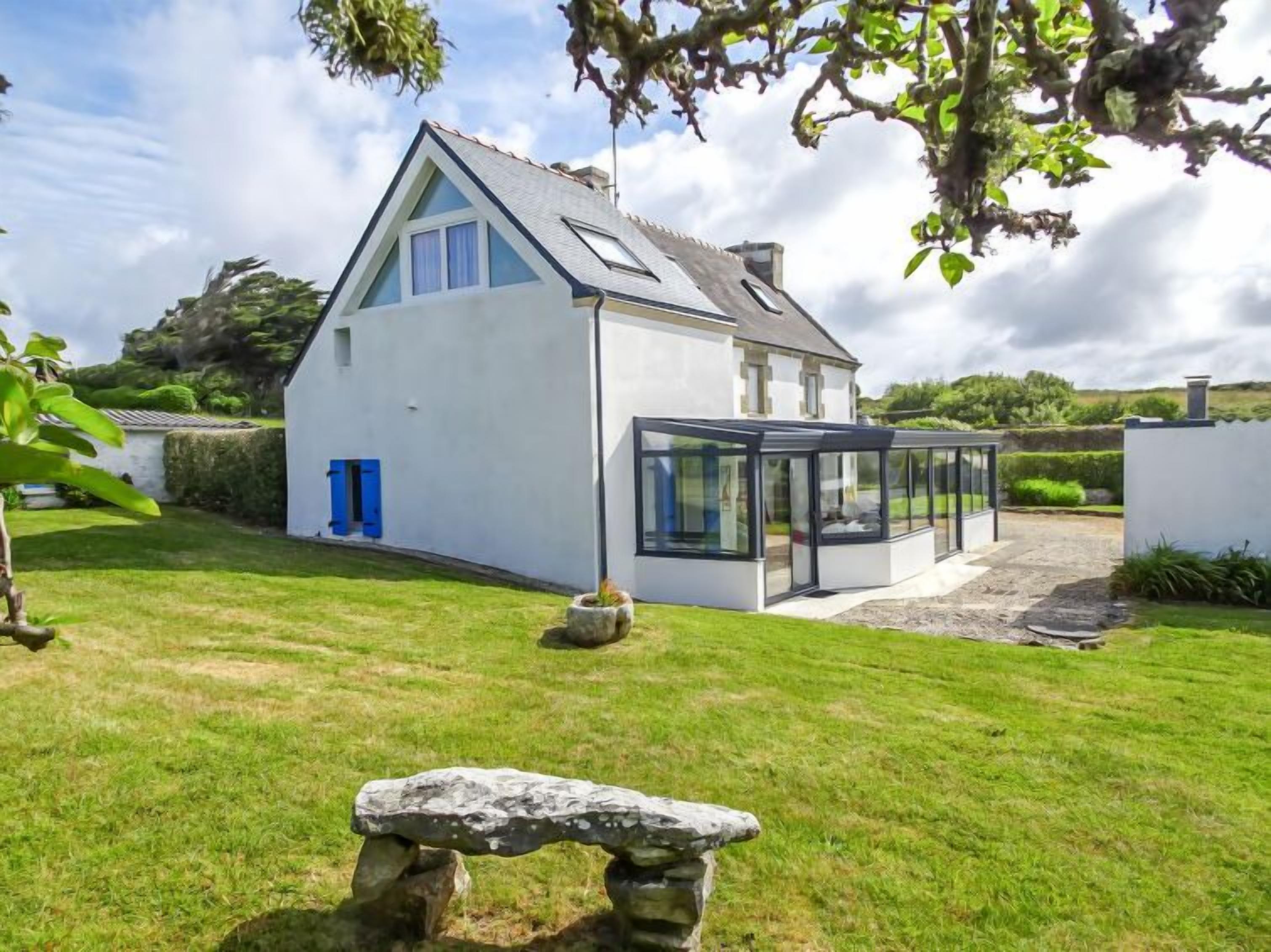 A white house with a triangular roof, a glass conservatory, and a stone bench in the yard.