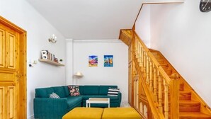Un salon avec un canapé de couleur turquoise, deux poufs de couleur moutarde, une petite table blanche et un escalier en bois.