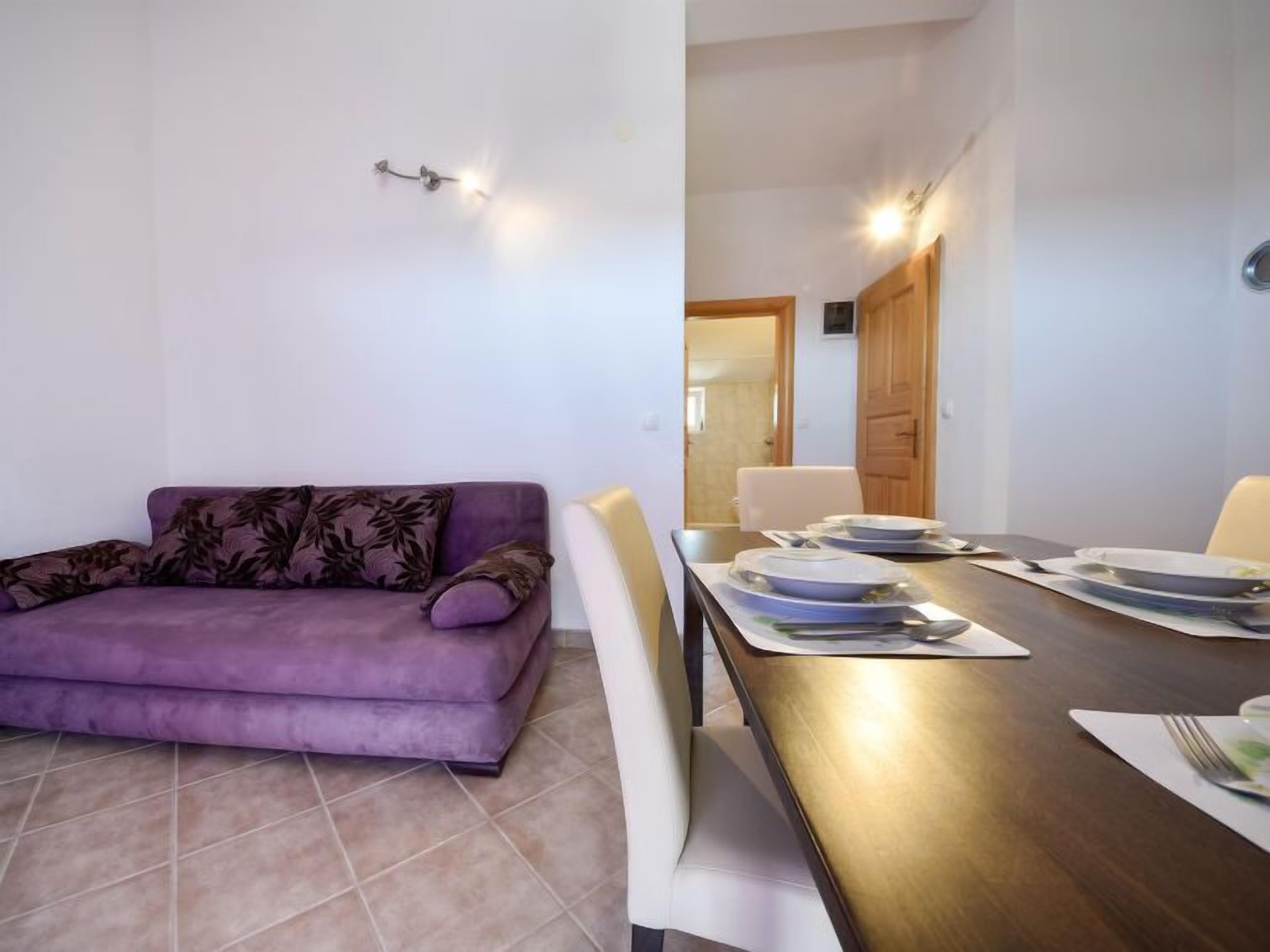A modern living room with a purple sofa, a dining table set with plates and cutlery, and a wooden door in the background.