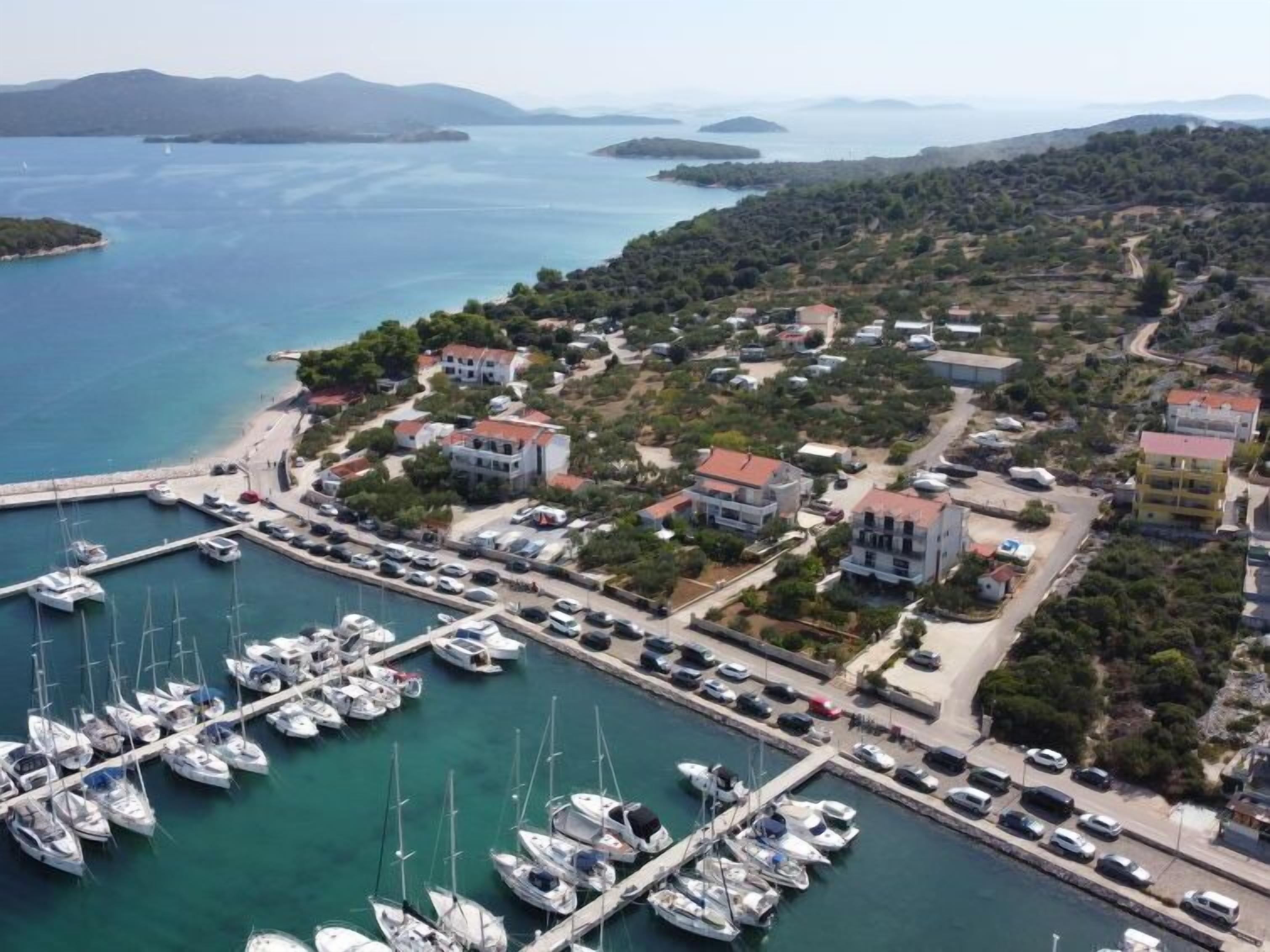 An aerial view of a coastal marina with multiple boats docked, surrounded by residential buildings and a hilly landscape in the background.
