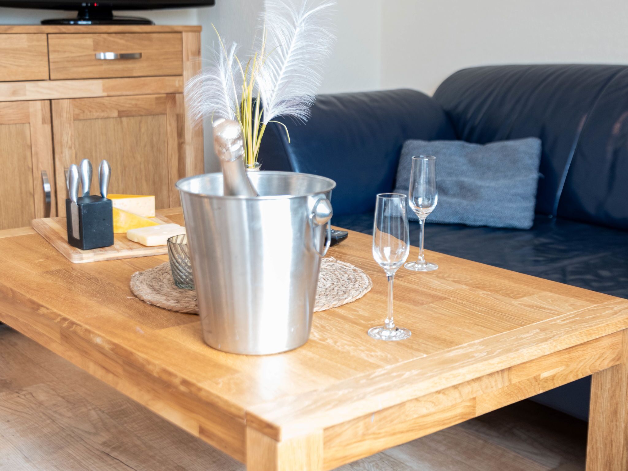 Una mesa de madera con un recipiente metálico, un bloque de cuchillos, un bloque de queso y dos copas de vino.