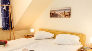 A neatly made bed with white linens and two rolled towels, a wooden headboard, a bedside table with a lamp, and a painting of a beach scene on the wall. - Strandläufer by Interhome (Norddeich)