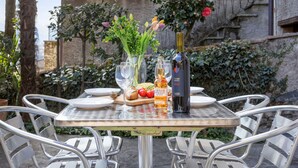 Mesa al aire libre con sillas de metal, una botella de vino, un vaso de agua, una hogaza de pan y una cesta pequeña de fruta.