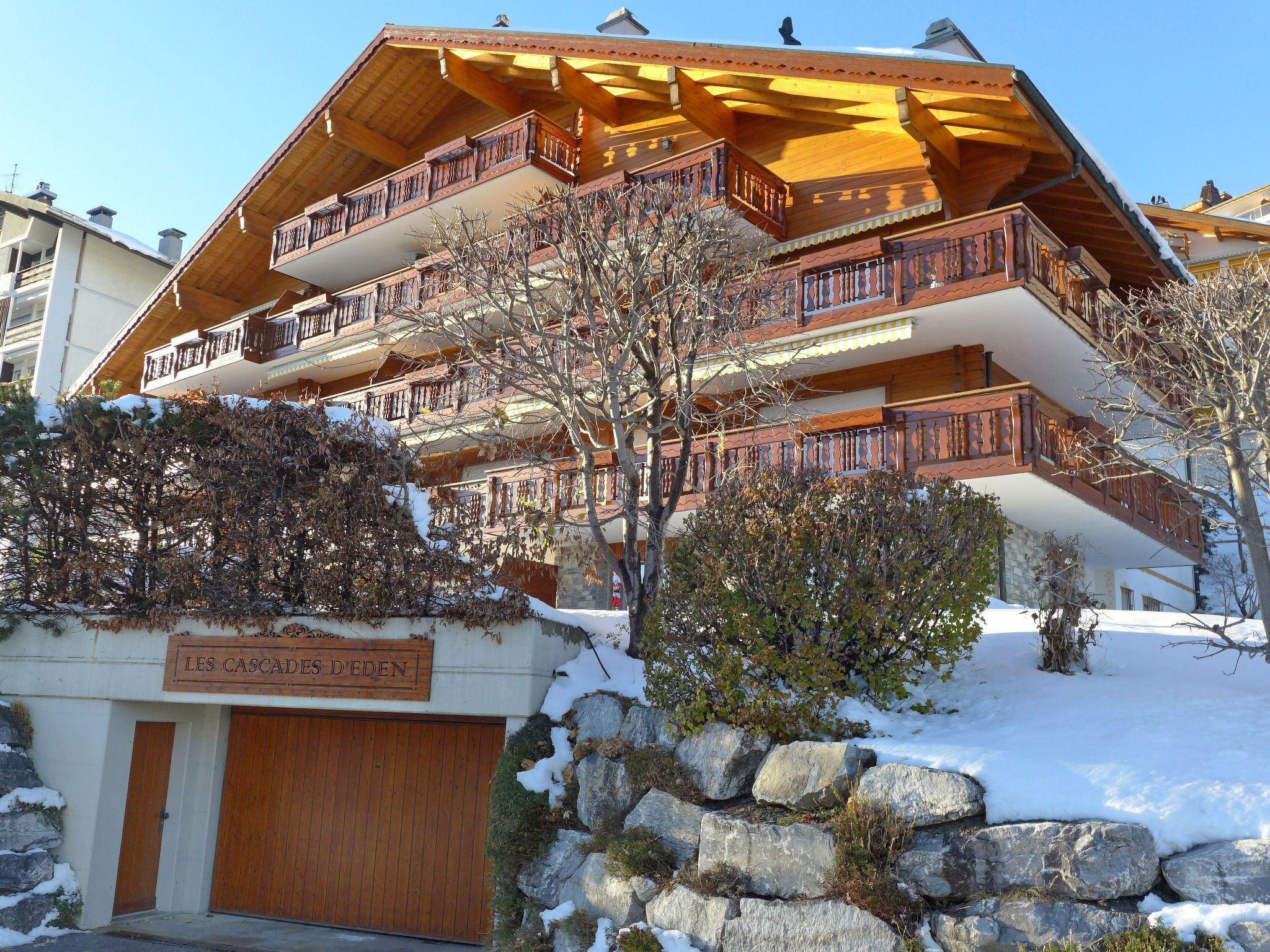 A multi-story wooden chalet with balconies, a stone wall, and a sign reading "LES CASCADES D'EDEN".