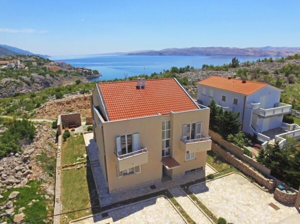A modern two-story house with a red-tiled roof, situated on a hillside with a view of a large body of water and mountains in the distance.