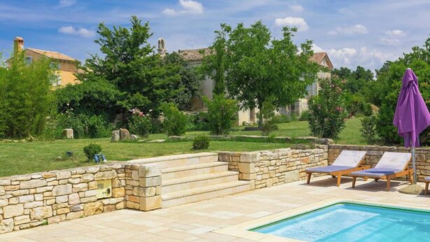 Area piscina con pareti in pietra, poltrone relax e un ombrellone viola.