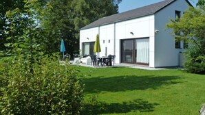 Een modern huis met twee verdiepingen, een witte gevel, een overdekt terras met tuinmeubilair en een verzorgd gazon.
