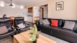Een moderne woonkamer met een zwarte leren bank, twee zwarte leren fauteuils, een houten salontafel met een plant, en een keuken op de achtergrond.