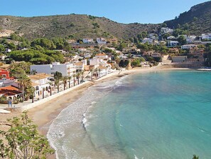 A coastal town with buildings along the beach, clear blue waters, and a hilly backdrop.