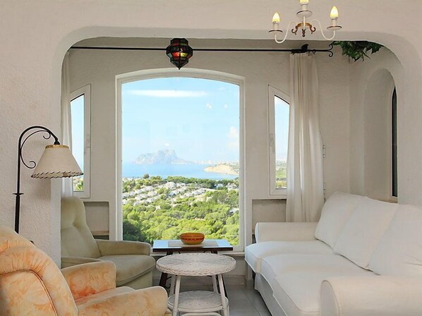 A room with a white sofa, a chair, a small table, and a view of a coastal landscape through an arched window.