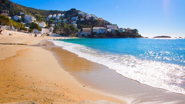 A coastal beach with clear blue waters, sandy shore, and buildings on a hillside. - Vacation home Bilbao by Interhome (Roses)