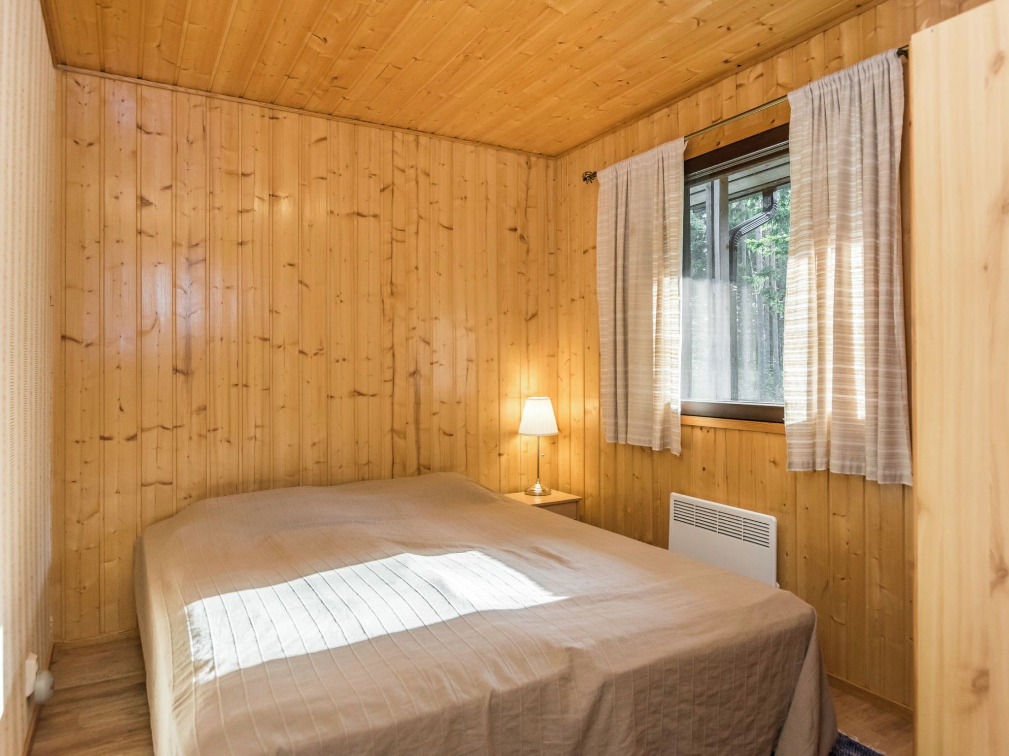 A wooden-paneled room with a bed, a window with curtains, and a wall-mounted heater.