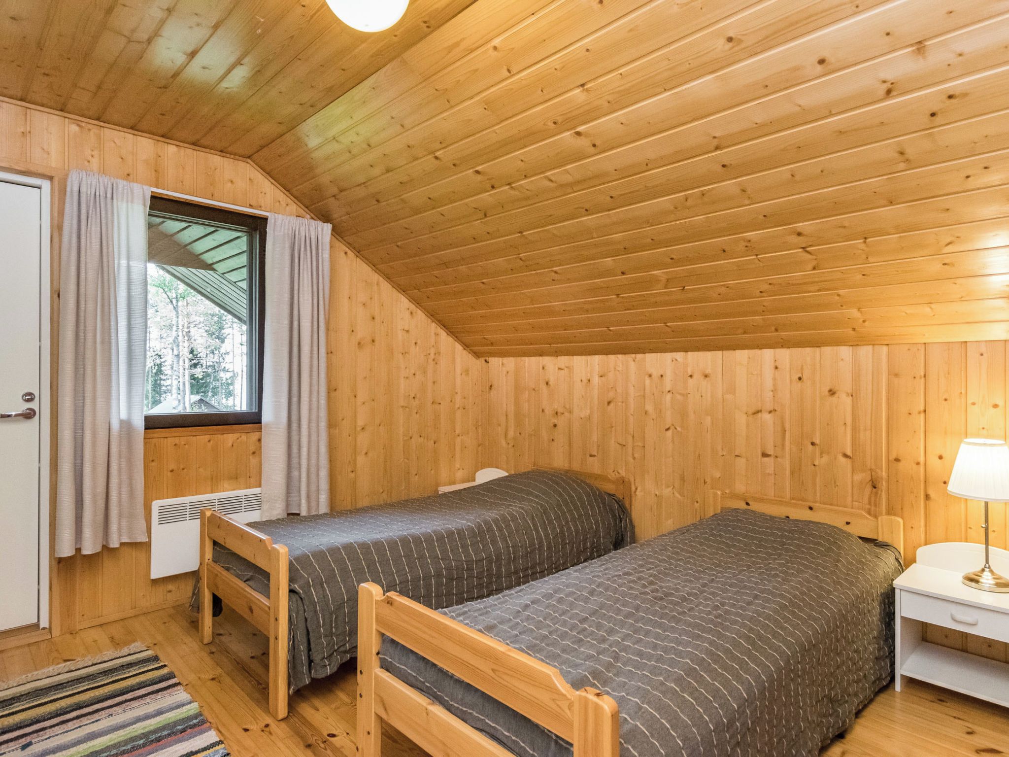 A small, cozy room with wooden walls and a sloped ceiling. There are two beds with matching bedding, a window with curtains, and a bedside table with a lamp.