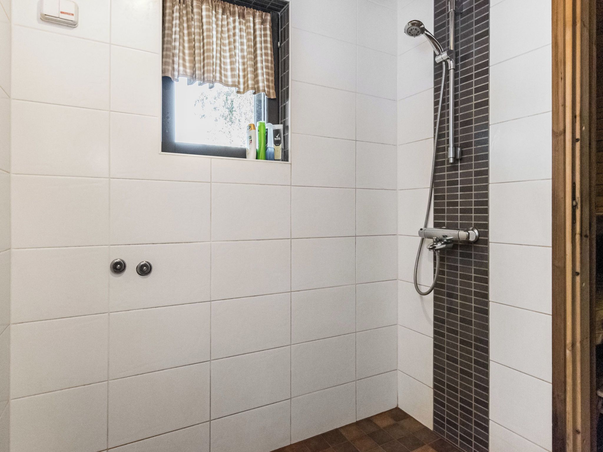 Une salle de bain avec des murs recouverts de carreaux blancs, une fenêtre ornée d’un rideau à carreaux, un espace douche avec un mur carrelé et un cadre de porte en bois.