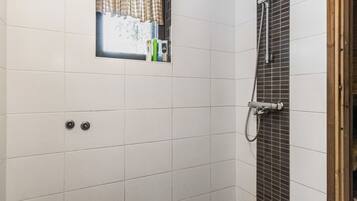 Une salle de bain avec des murs recouverts de carreaux blancs, une fenêtre ornée d’un rideau à carreaux, un espace douche avec un mur carrelé et un cadre de porte en bois.