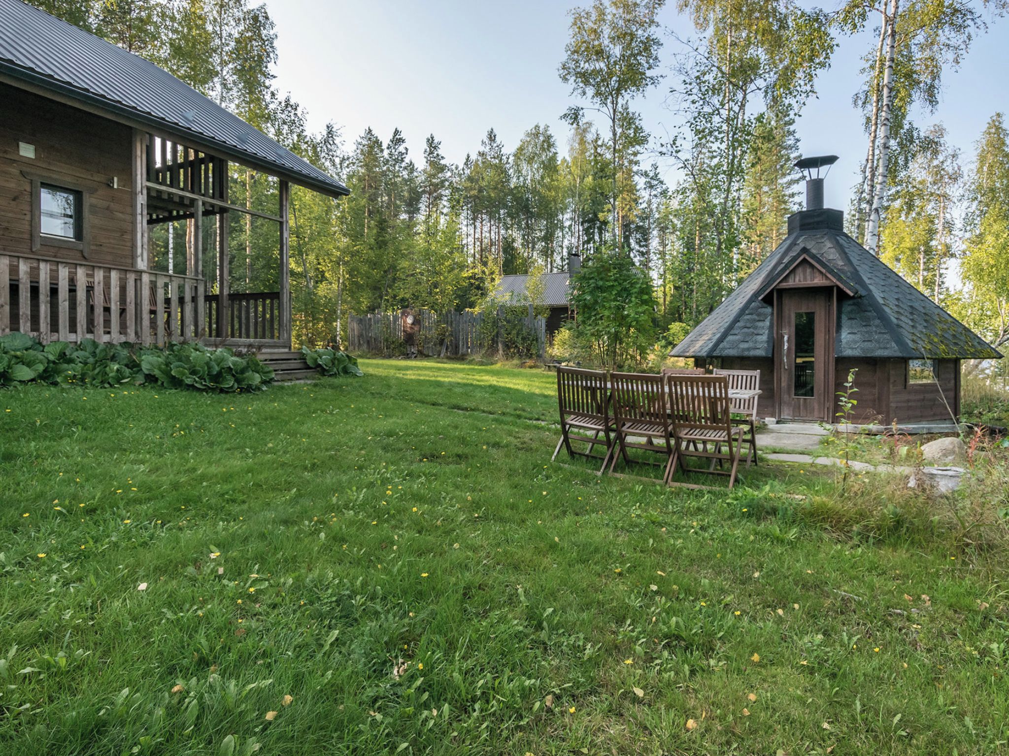 A rustic cabin with a wooden deck and a chimney, surrounded by a grassy yard and dense trees.