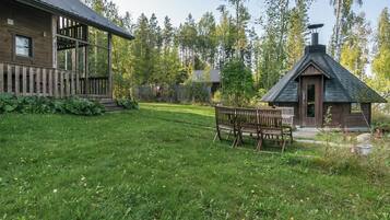 A rustic cabin with a wooden deck and a chimney, surrounded by a grassy yard and dense trees.