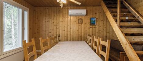 Een kamer met houten lambrisering, een lange tafel, stoelen, een plafondventilator, een airconditioner, een klok en een trap.