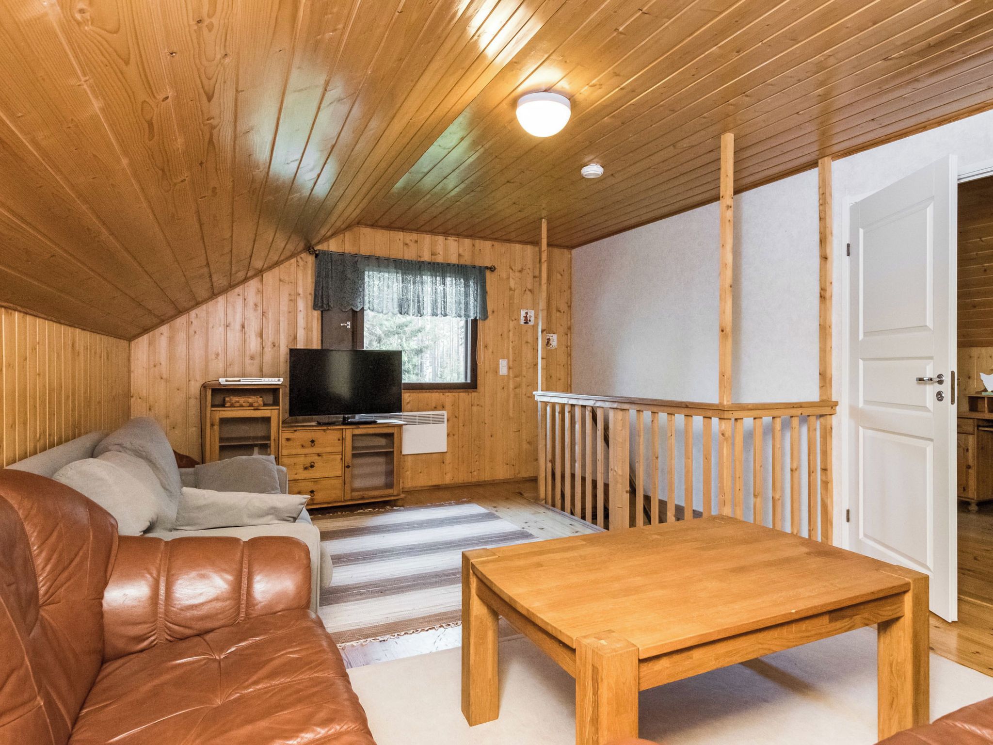 A cozy cabin interior with a wooden ceiling, a television, a sofa, a table, and a window with curtains.