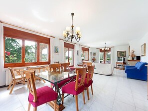 A spacious living area with a glass dining table surrounded by wooden chairs and pink cushions, a blue sofa, and a television in the background.