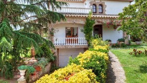 Une maison à deux étages dotée d’un balcon, entourée d’une végétation luxuriante et de plantes en fleurs.