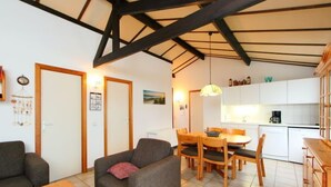 Een gezellige woonkamer met een bank, een houten eettafel en een keukenhoek met witte kasten en een hanglamp.