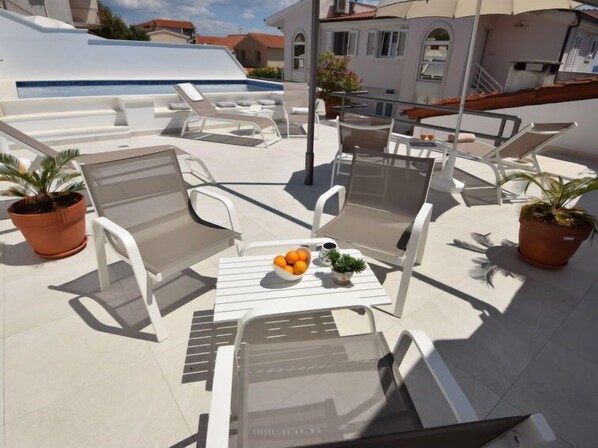 Une terrasse sur le toit, avec des chaises longues blanches, une table sur laquelle se trouve un saladier rempli d’oranges et une petite plante, ainsi que des plantes en pot.