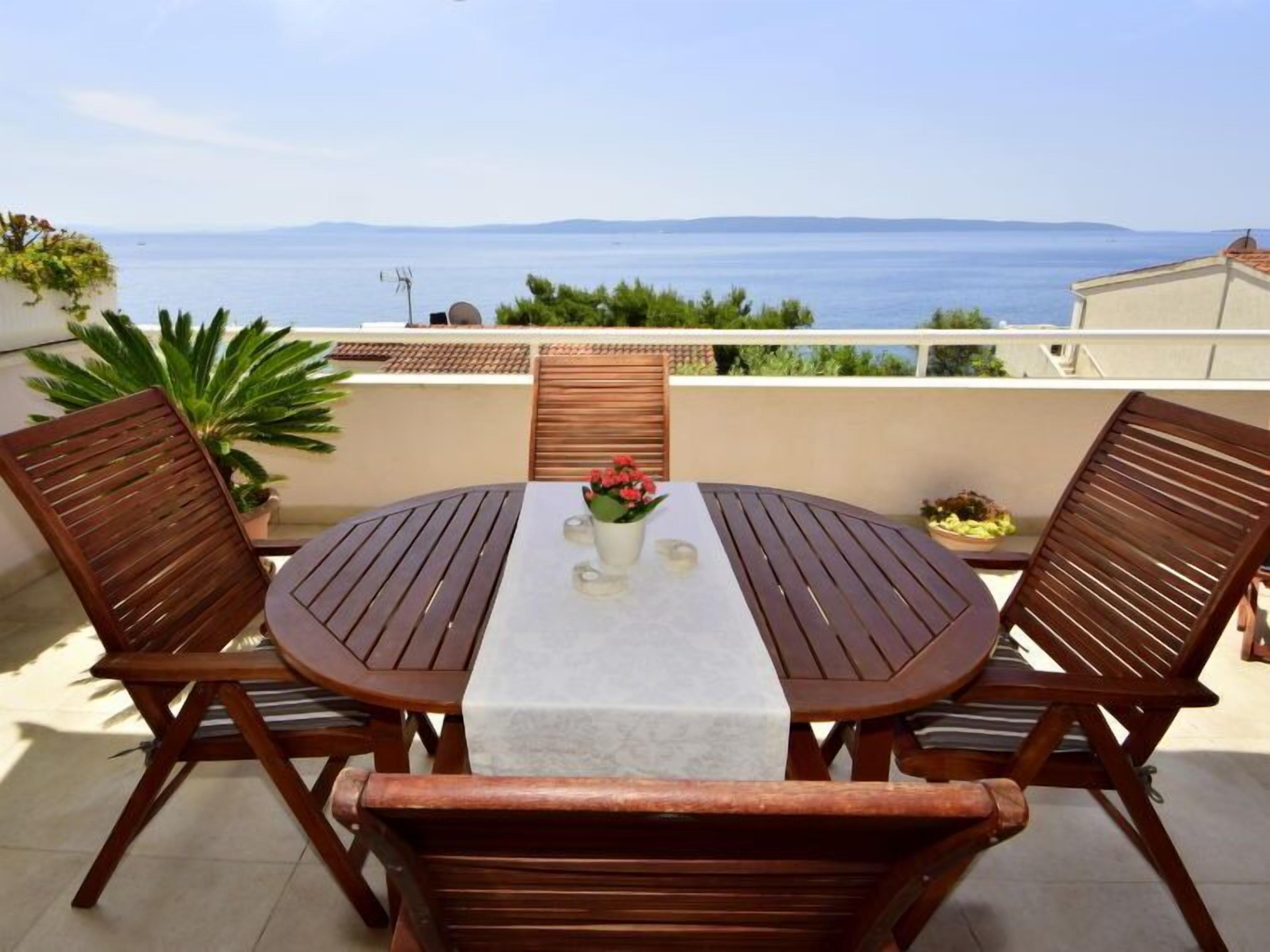 Une table ronde recouverte d’une nappe blanche, entourée de quatre chaises en bois, est placée sur un balcon offrant une vue sur la mer.