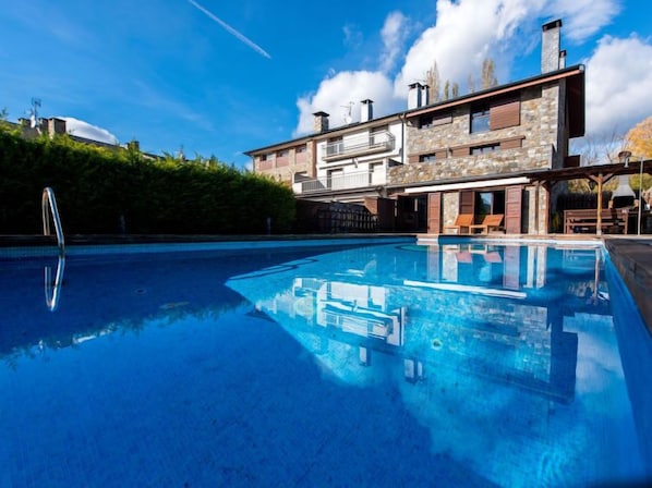 A modern poolside area with a clear blue pool, a stone facade building, and a well-maintained hedge.
