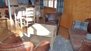 Een gezellige woonkamer met een houten salontafel, leren fauteuils en een bank. Achtergrond toont een eethoek met een houten tafel en stoelen.