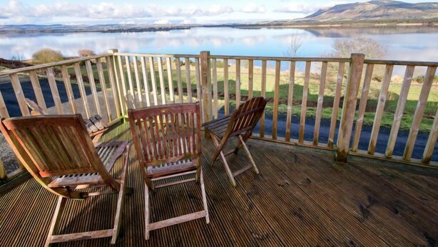 Une terrasse en bois avec trois chaises en bois, donnant sur un lac et entourée d’une balustrade en bois.
