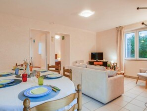 A well-lit living room with a dining area set for four, a sofa, a TV, and a window with curtains. - Mer et Soleil by Interhome (Carnac)