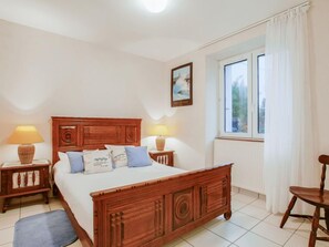 A bedroom with a wooden bed, two bedside tables with lamps, a chair, a window with curtains, and a painting on the wall. - Mer et Soleil by Interhome (Carnac)