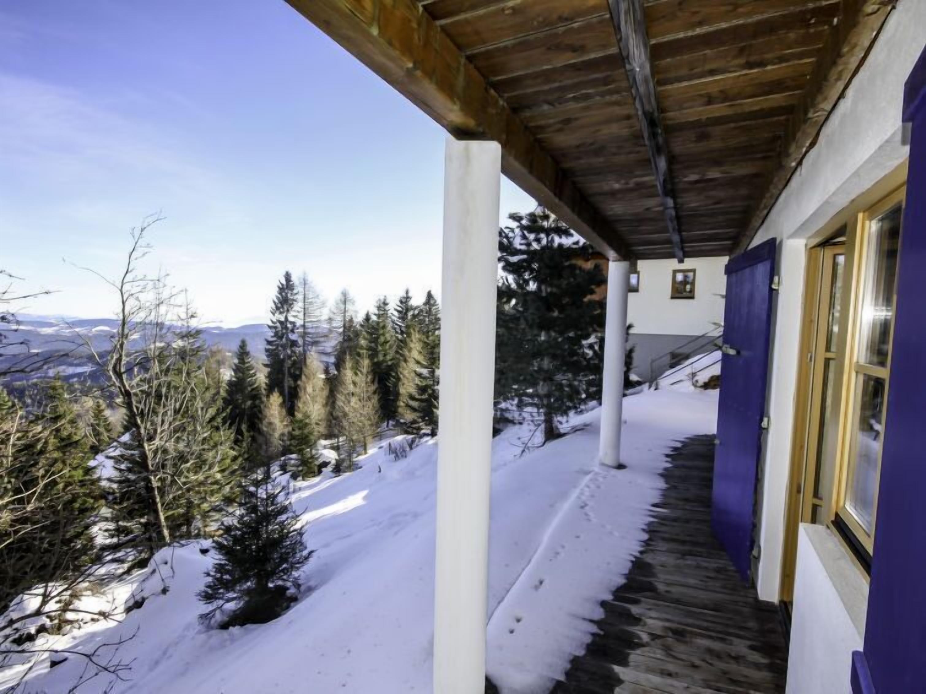 A covered outdoor area with a wooden ceiling, a snow-covered path leading to a building with a purple door, and a view of a snowy landscape with trees and distant mountains.
