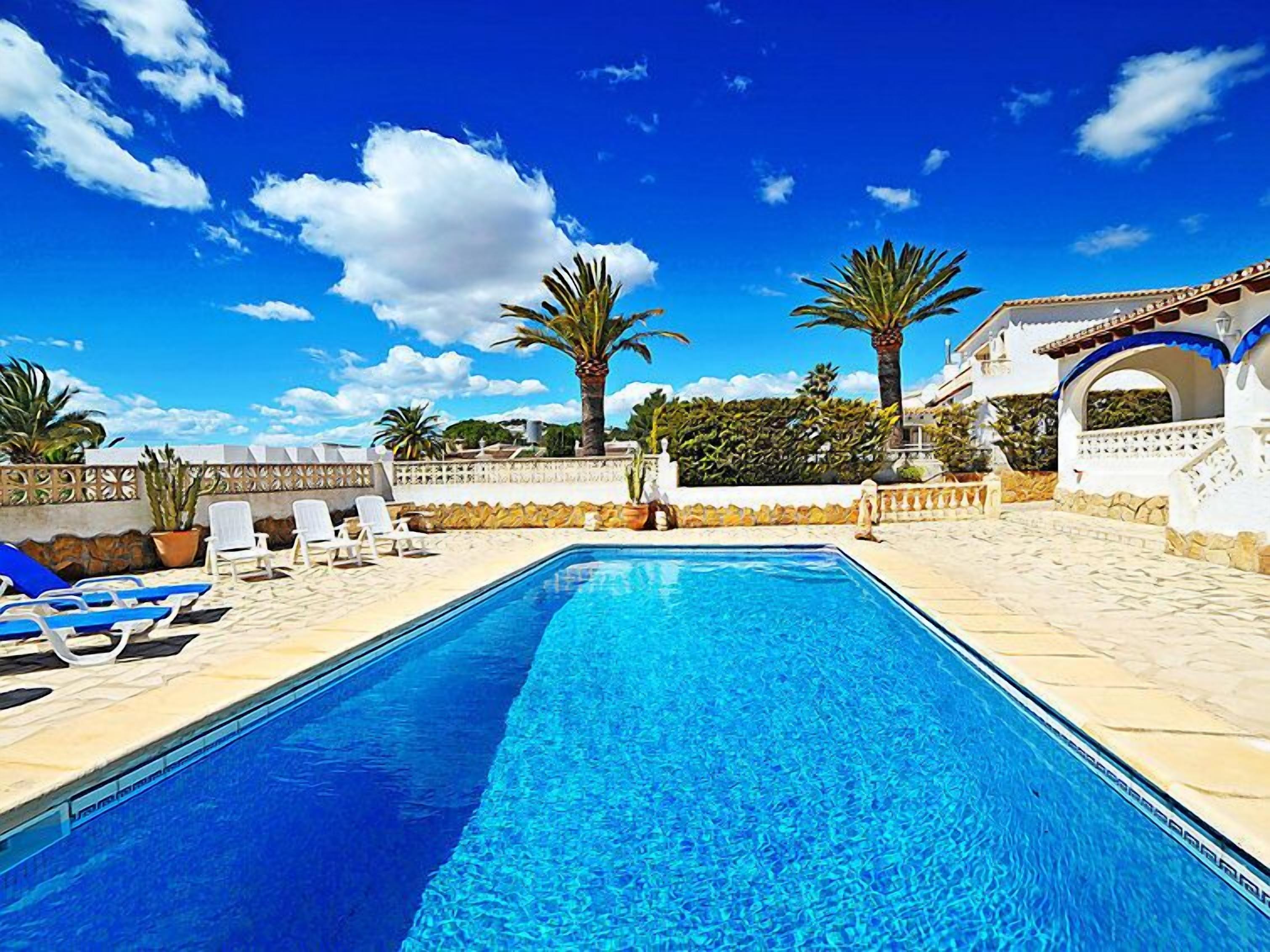 A rectangular swimming pool with clear blue water, surrounded by white lounge chairs, palm trees, and a white building with a blue roof.