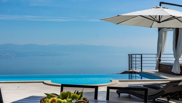 A poolside area with a view of the sea, a wooden table, a wicker basket, and a white umbrella.